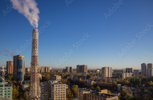 A beautiful urban landscape with tall modern houses and a metal pipe of a thermal power plant against the background of a clear blue sky on an autumn day