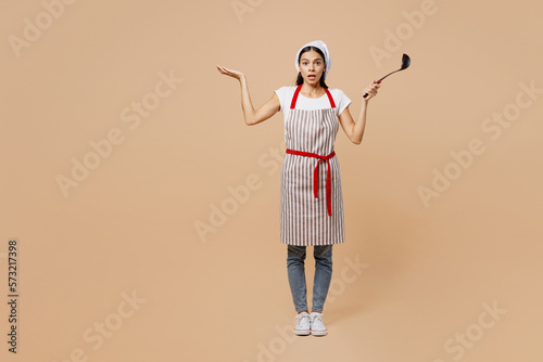 Full body confused shocked young housewife housekeeper chef baker latin woman wear apron toque hat hold in hand laddle spread hand isolated on plain pastel light beige background Cook food concept