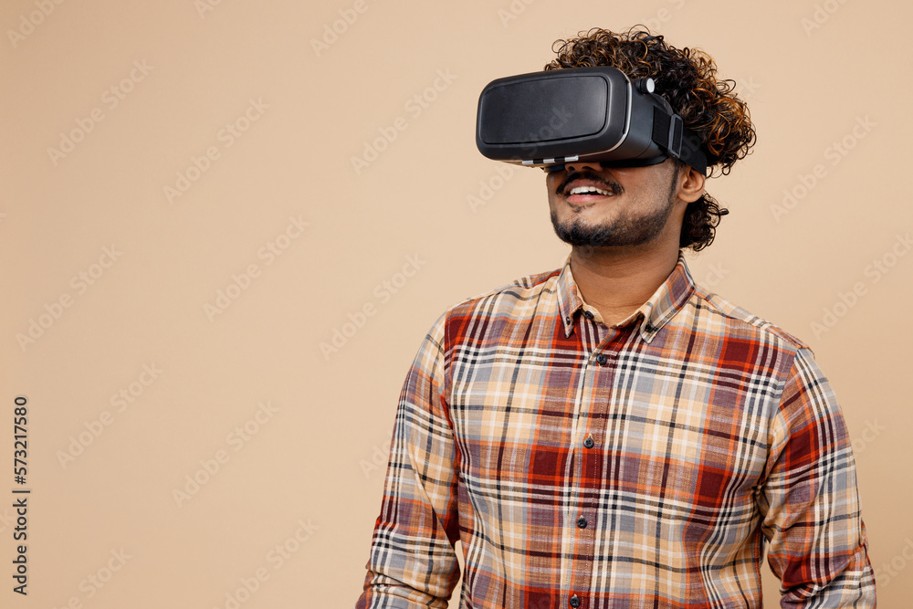 Young smiling cheerful gambling Indian man wear brown shirt casual clothes watching in vr headset pc gadget isolated on plain pastel light beige background studio portrait. People lifestyle concept. Young smiling cheerful gambling Indian man wear brown shirt casual clothes watching in vr headset pc gadget isolated on plain pastel light beige background studio portrait. People lifestyle concept.