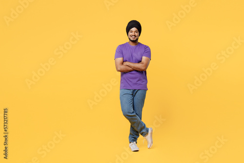 Full body smiling happy fun devotee Sikh Indian man ties his traditional turban dastar wear purple t-shirt hold hands crossed folded look camera isolated on plain yellow background studio portrait.
