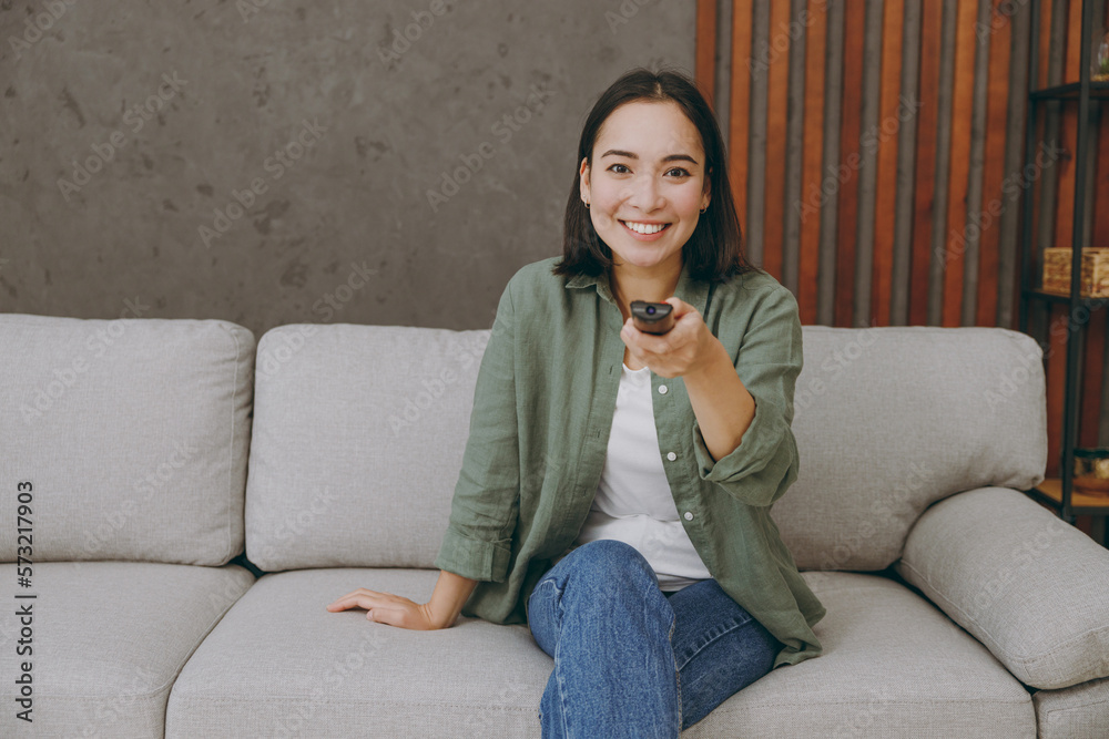 Young smiling happy fun woman of Asian ethnicity wears casual clothes watch TV switch channels sits on grey sofa couch stay at home hotel flat rest relax spend free spare time in living room indoor. Young smiling happy fun woman of Asian ethnicity wears casual clothes watch TV switch channels sits on grey sofa couch stay at home hotel flat rest relax spend free spare time in living room indoor.
