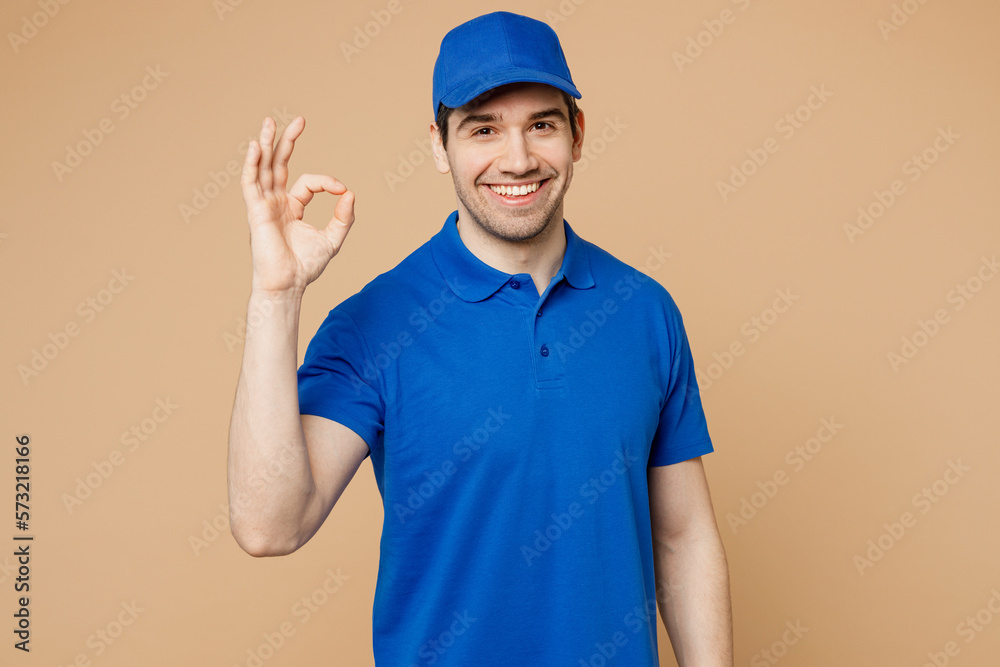 Professional satisfied smiling delivery guy employee man wear blue cap t-shirt uniform workwear work as dealer courier showing okay ok gesture isolated on plain light beige background Service concept