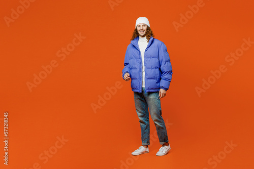 Full body smiling happy fun cheerful young man with long curly hair wear hat purple ski padded jacket casual clothes walking going look camera isolated on plain orange red background studio portrait.