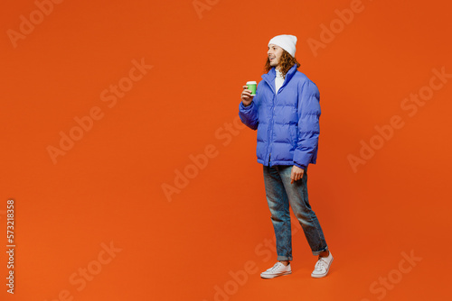 Full body side view young man with long curly hair wear hat purple ski padded jacket casual clothes hold takeaway delivery craft paper brown cup coffee to go isolated on plain orange red background.