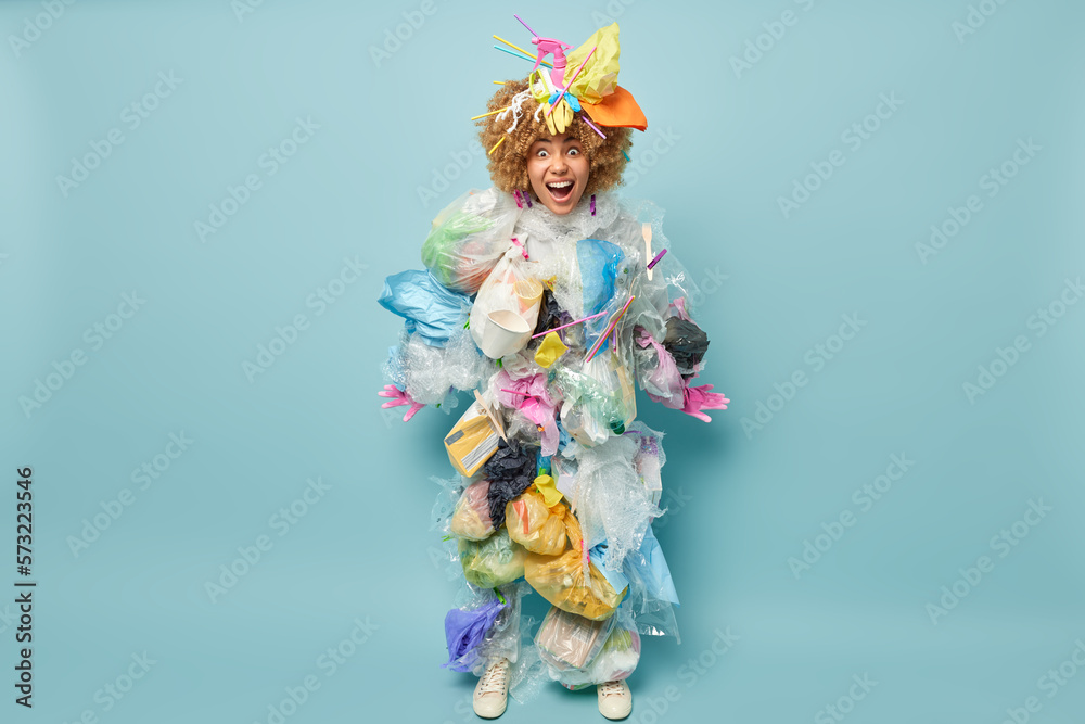Full length shot of curly haired female volunteer poses in clothes of garbage stares with widely opened eyes cannot believe own eyes isolated over blue background. Recycling and environmental concept