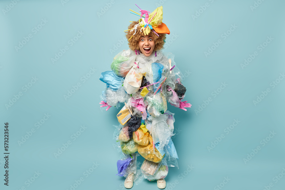 Fototapeta premium Full length shot of curly haired female volunteer poses in clothes of garbage stares with widely opened eyes cannot believe own eyes isolated over blue background. Recycling and environmental concept