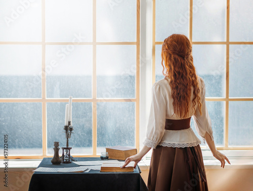 mysterious redhead woman stands in room near window waiting for love. Girl writer looking for inspiration. Long flowing red hair back rear view. lady queen vintage dress. royal cabinet old style room photo