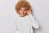 Portrait of cheerful curly woman smiles and looks happily expresses candid joy keeps hand on rim of spectacles wears casual long sleeved jumper isolated on white background. Positive emotions concept