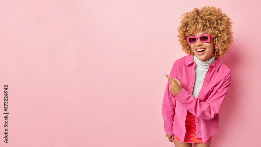 Overjoyed stylish woman wears sunglasses shirt and skirt points finger ...