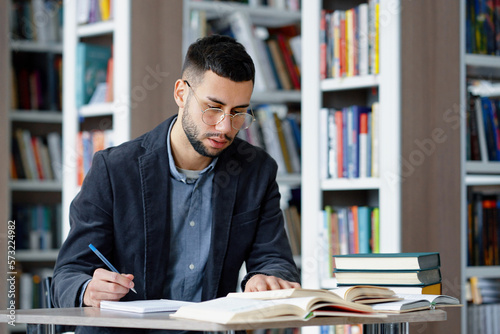 Student wearing eyeglasses writing homework in library, speaking aloud his ideas, heap of books lying beside, bookcases on blurred background. Concept of study