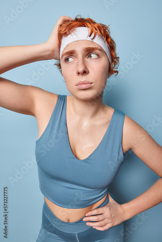 Sweaty fitness woman stands tired after doing physical exercises keeps hand on waist focused aside unhappily wears white headband and tracksuit isolated over blue background. Sporty lifestyle