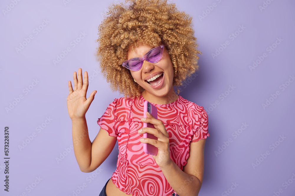 Waist up shot of happy overjoyed woman has curly hair keeps palm raised holds phone near mouth sings song wears purple sunglasses pink tshirt poses indoor has fun. Carefree optimistic female model Waist up shot of happy overjoyed woman has curly hair keeps palm raised holds phone near mouth sings song wears purple sunglasses pink tshirt poses indoor has fun. Carefree optimistic female model