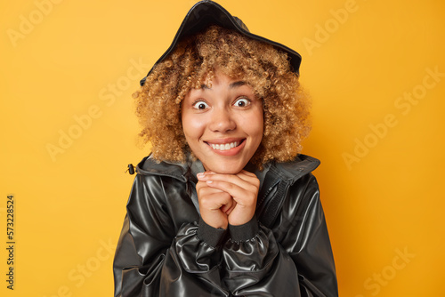 Portrait of cheerful surprised woman with curly hair keeps hands under chin bites lips cannot believe in something unexpected wears black leather jacket and hat isolated over yellow background photo