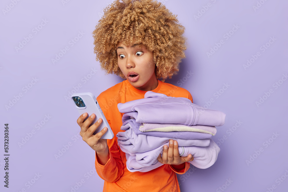 Startled curly haired European woman stares with omg expression at camera holds neatly folded washed clothes cannot believe own eyes dressed in orange jumper isolated over purple background. Startled curly haired European woman stares with omg expression at camera holds neatly folded washed clothes cannot believe own eyes dressed in orange jumper isolated over purple background.