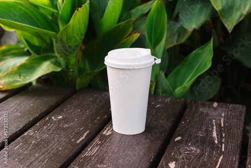 Takeaway paper cup standing on wood table with tropical palm leaves background. Travel, eco, coffee to go, summer vacation concept. Mock up, mockup, blank, place for logo or text
