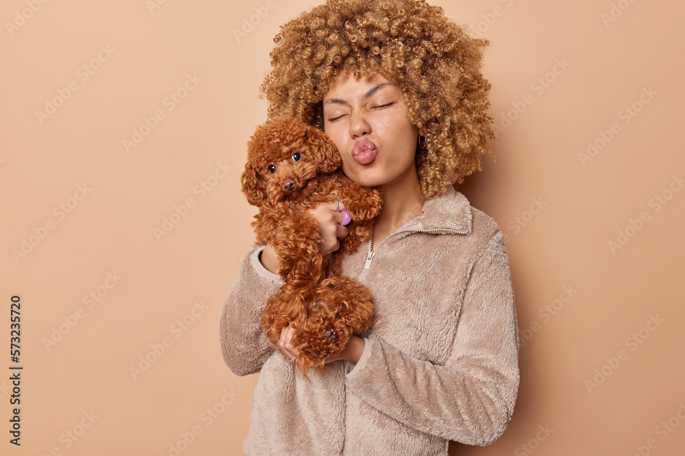 Curly haired young woman poses with cute puppy keeps lips folded dressed in fur jacket poses with affectionate poodle dog isolated over brown background. Domestic animal owner poses indoors. Curly haired young woman poses with cute puppy keeps lips folded dressed in fur jacket poses with affectionate poodle dog isolated over brown background. Domestic animal owner poses indoors.