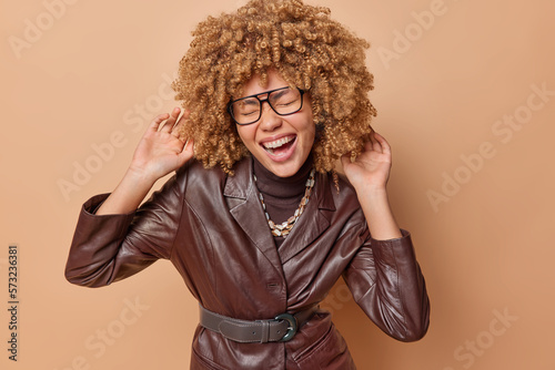 Overjoyed curly haired woman exclaims loudly wears transparent eyeglasses and leather jacket feels very happy smiles broadly shows white teeth isolated over brown background. Positive emotions concept © Wayhome Studio