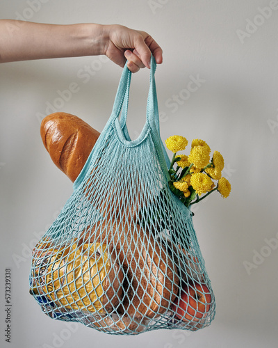 the hand holds a mesh bag (string bag) with products. bread, flowers, loaf, apple and fruit inside