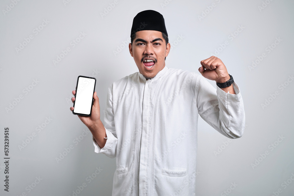 Excited young Asian Muslim man showing mobile phone screen and celebrating success isolated on white background. Excited young Asian Muslim man showing mobile phone screen and celebrating success isolated on white background.