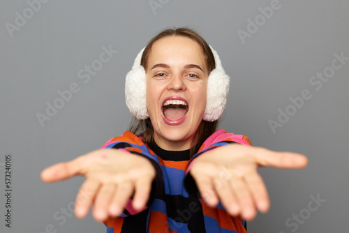 Extremely happy woman with brown hair wearing jumper and warm earmuffs standing isolated over gray background, rejoicing good news, sharing her good mood with you, posing with outstretched hands.