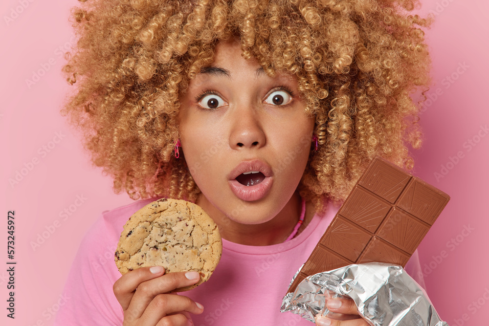 Close up shot of stunned woman holds breath from amazement keeps mouth opened holds cookie and bar of chocolate eats unhealthy sweet food isolated over pink background. Food addiction concept