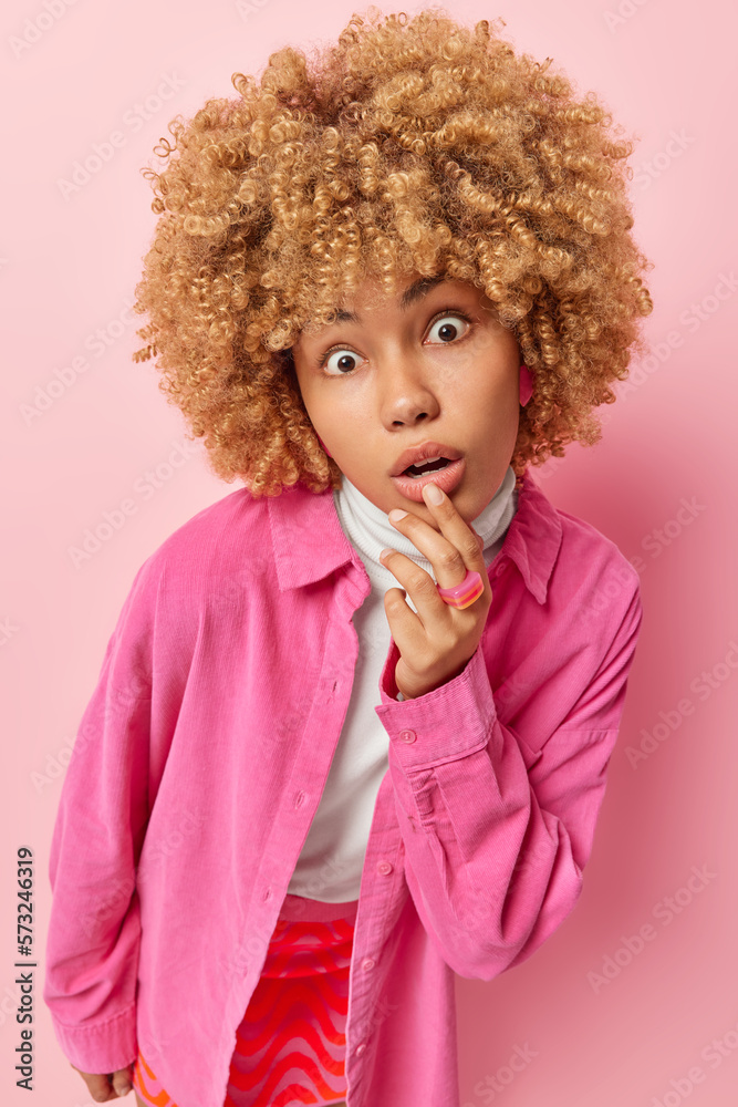 Vertical shot of wondered woman with curly hair keeps mouth opened from shock looks curious wears stylish shirt and skirt has widely opened eyes poses against pink background. Amazement concept Vertical shot of wondered woman with curly hair keeps mouth opened from shock looks curious wears stylish shirt and skirt has widely opened eyes poses against pink background. Amazement concept