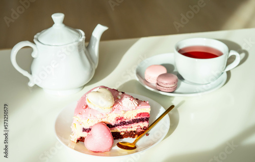Pink cake with heart shaped macaroons , red tea, white kettle and gold spoon on a black background. Natural light, sunny day