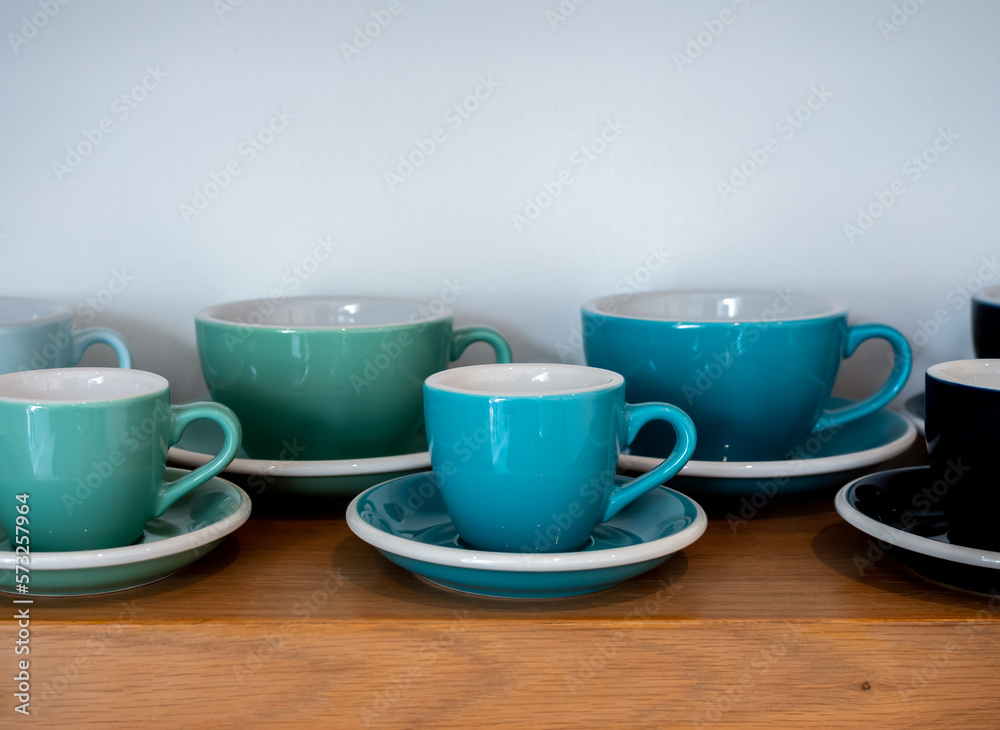 Close-up set of big and small blue green ton of coffee mugs, ceramic cups with saucers stand in a row on wooden shelf isolated on white background with copy space. Close-up set of big and small blue green ton of coffee mugs, ceramic cups with saucers stand in a row on wooden shelf isolated on white background with copy space.