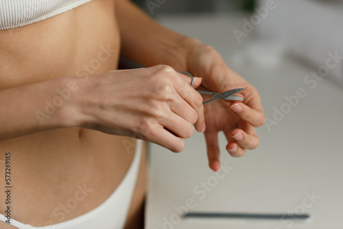 Young woman in underwear is standing in the bathroom and trimming her nails with nail scissors. Hygienic procedures, healthy body care. Close-up of slender figure. Girl uses small scissors for nails.