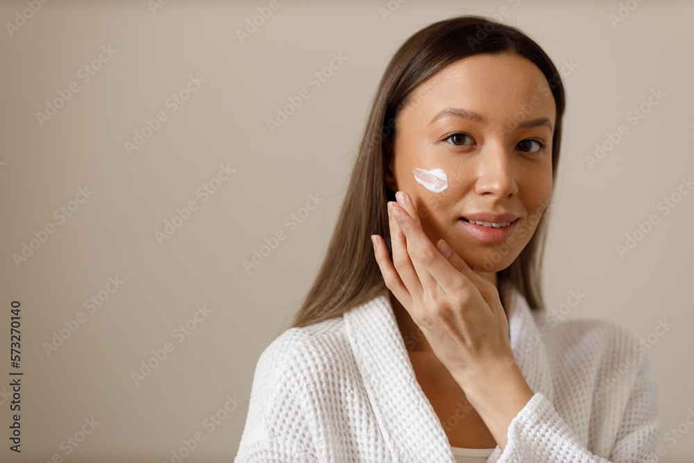 Attractive young woman in white bathrobe applies lotion or moisturizer to her face. Professional facial care after washing. Portrait of brunette girl on isolated background looking into the camera. Attractive young woman in white bathrobe applies lotion or moisturizer to her face. Professional facial care after washing. Portrait of brunette girl on isolated background looking into the camera.