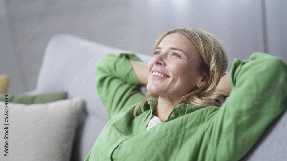 Close up of happy mature blonde woman relaxing sitting on sofa in living room at home. Smiling female in casual clothes stretches with hands behind her head and eyes closed indoors. Has a good mood Close up of happy mature blonde woman relaxing sitting on sofa in living room at home. Smiling female in casual clothes stretches with hands behind her head and eyes closed indoors. Has a good mood