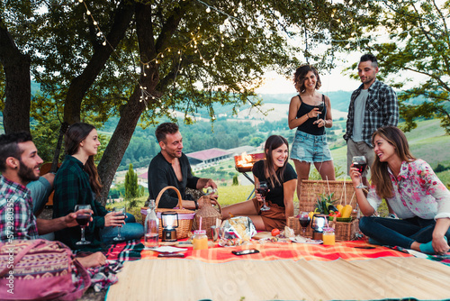 Group of happy friends having fun at the picnic barbecue in a countryside area, celebrating, eating grilled food and drinking tasty wine