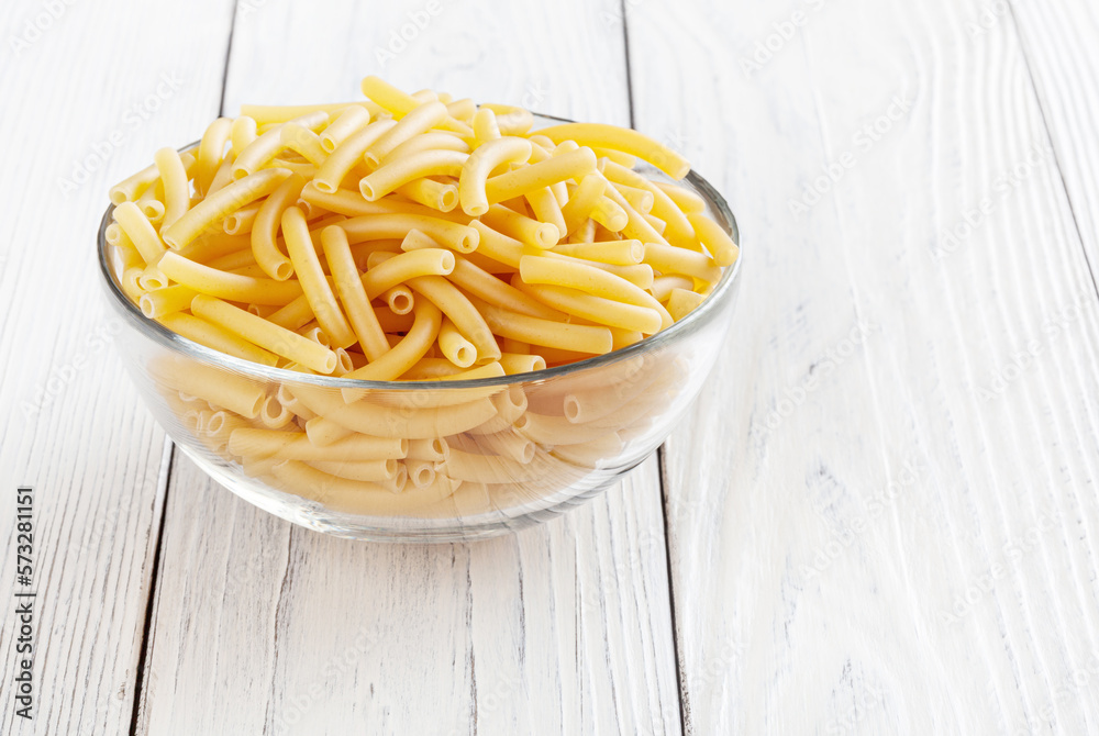 Uncooked maccheroni pasta in glass bowl on white wooden background