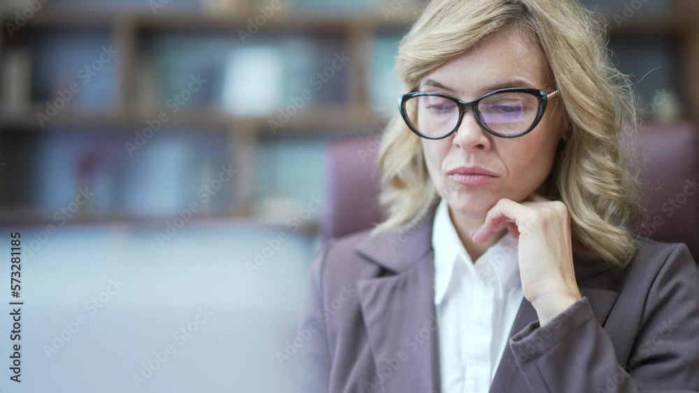 Close up of thoughtful mature businesswoman in suit and glasses looking seriously at laptop screen at desk in office. Concentrated confident female owner is thinking about solving a complex problem Close up of thoughtful mature businesswoman in suit and glasses looking seriously at laptop screen at desk in office. Concentrated confident female owner is thinking about solving a complex problem
