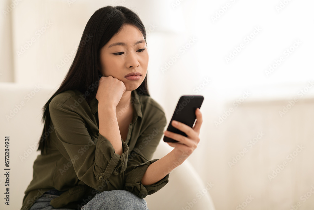 Bored asian lady sitting on couch at home, using smartphone Bored asian lady sitting on couch at home, using smartphone