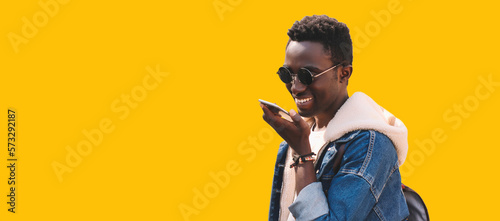 Portrait of happy smiling young african man holding smartphone using voice command recorder, assistant or takes calling, looking at phone isolated on yellow background