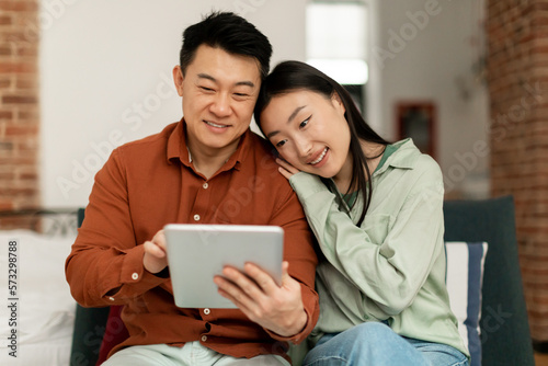 Happy korean spouses using digital pad, surfing internet or shopping online, sitting on sofa at home, free space