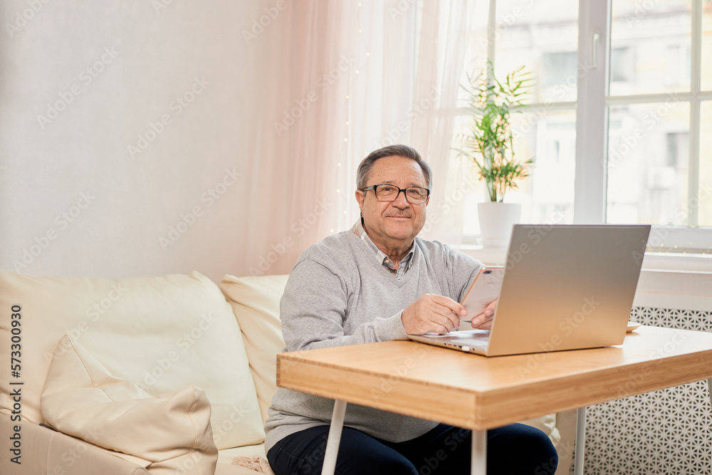 Cheerful senior man has phone conversation, chatting on the smartphone working with a laptop remotely from home. Cheerful senior man has phone conversation, chatting on the smartphone working with a laptop remotely from home.