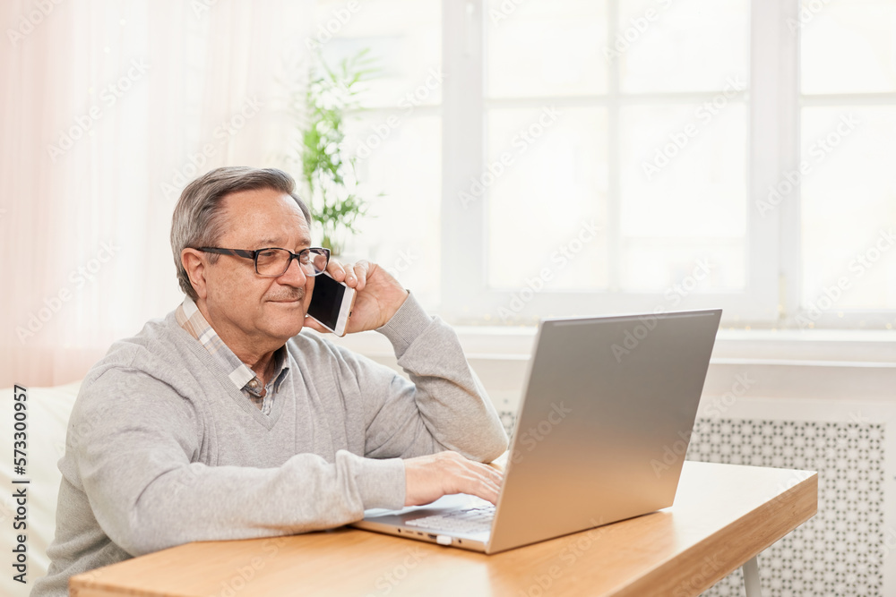 Cheerful senior man has phone conversation, chatting on the smartphone working with a laptop remotely from home. Cheerful senior man has phone conversation, chatting on the smartphone working with a laptop remotely from home.