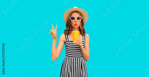 Summer portrait of beautiful young woman drinking fresh juice wearing straw round hat, striped dress on blue background