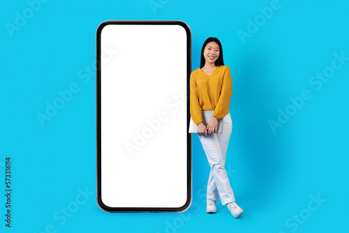 Cool asian lady with laptop standing by big phone, mockup