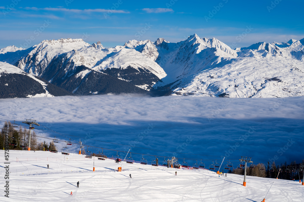 People skiing on ski slope and going up the hill on chair ski lift. Inversion clouds in a valley. High altitude ski resort La Plagne, Savoie, France