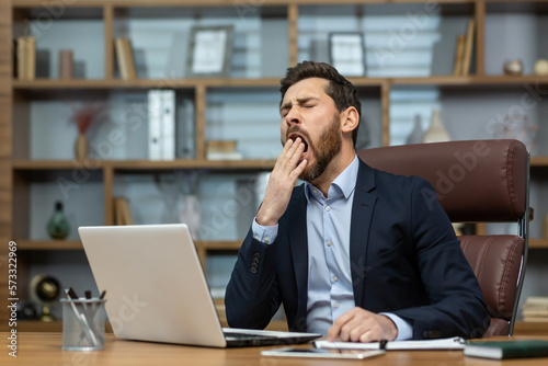 Mature man yawning inside classic office, senior businessman in workplace sleepless and overworked, boss in business suit sitting at desk, using laptop at work.
