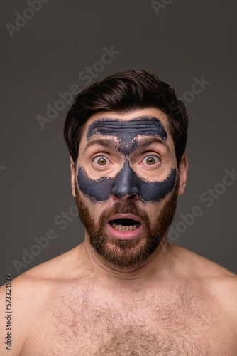 Shocked shirtless man with clay mask on face looking at camera isolated on grey background 