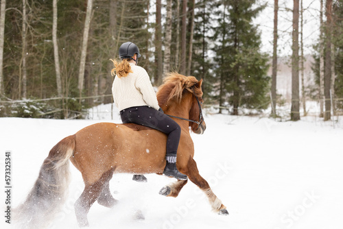 Icelandic horse and female rider riding on snowy field. Rider has black helmet. © AnttiJussi