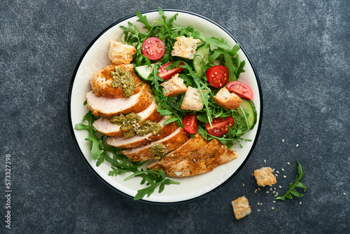 Fresh salad with grilled chicken fillet, lettuce, arugula, spinach, tomatoes, cucumber and pesto sauce, sesame seeds with olive oil in white bowl on black slate background. Healthy lunch menu Top view