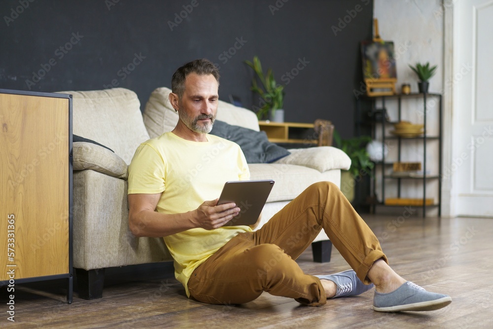 Middle-age beard male student wearing casual dress sitting on floor in the living room and studying online, using digital tablet. Communicating with friends.