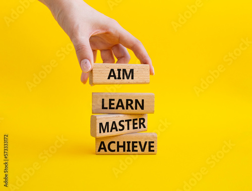 Aim Learn Master Achieve symbol. Concept word Aim Learn Master Achieve on wooden blocks. Beautiful yellow background. Businessman hand. Business and Aim Learn Master Achieve concept. Copy space © Natallia