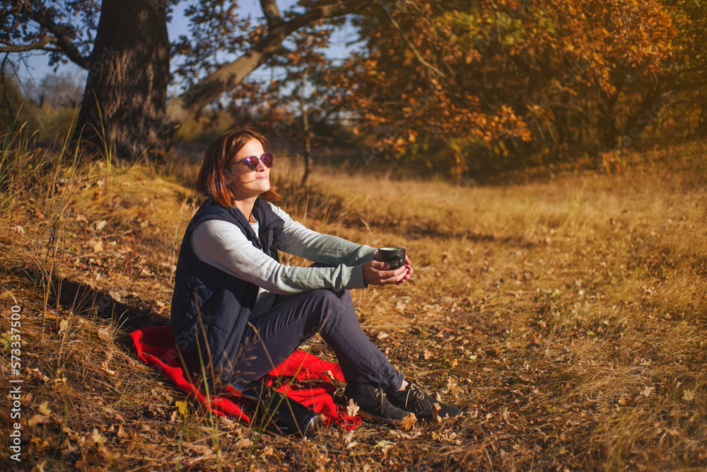 A young girl in an autumn park is resting on the grass with a cup of tea in her hands on a sunny warm day against the backdrop of autumn trees A young girl in an autumn park is resting on the grass with a cup of tea in her hands on a sunny warm day against the backdrop of autumn trees