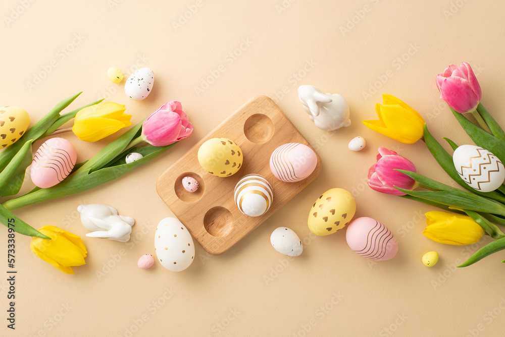 Easter concept. Top view photo of colorful easter eggs in wooden holder ceramic easter bunnies yellow and pink tulips on isolated pastel beige background